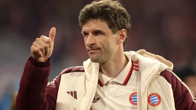 Thomas Müller, com a jaqueta bege e vinho do Bayern de Munique, em campo, acenando com a mão