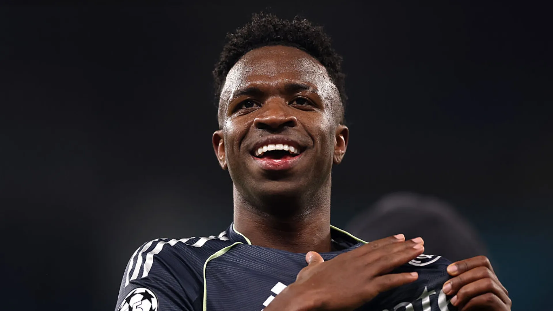 Vini Jr. com a mão no escudo do Real Madrid, enquanto sorri